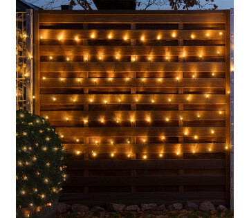Een hek en een bosjes zijn versierd met de netverlichting. De lampjes geven warm wit licht. 