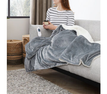 Een vrouw zit onder het SleepMed Elektrische Knuffeldeken in de lichtgrijs. 